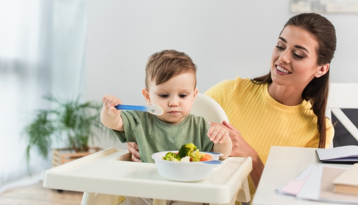 Smiling mother looking at son with spoon near vegetables in bowl on high chair.