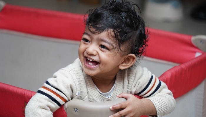 A closeup photo of an adorable indian toddler baby boy smiling with dimple in cheeks and standing inside a playpen.