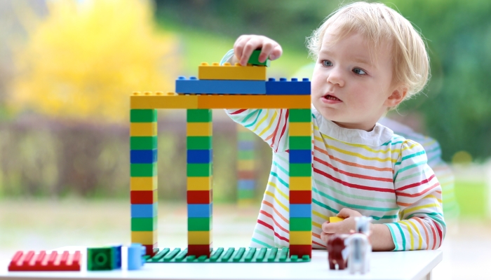 Cute blonde toddler girl building house from plastic blocks.