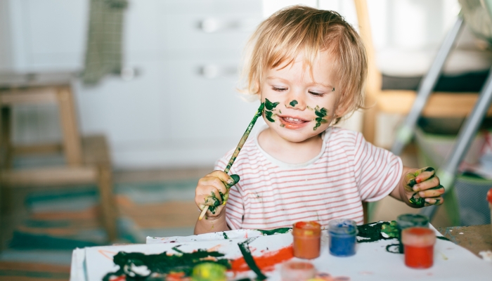 Happy cute toddler painting her face with gouache paints.