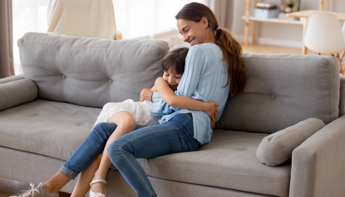 Happy mother hugging cute little girl sitting together on sofa in living room.