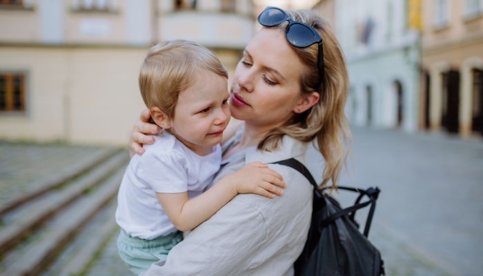 Mother consling her little daughter crying, holding her in arms in street in summer.