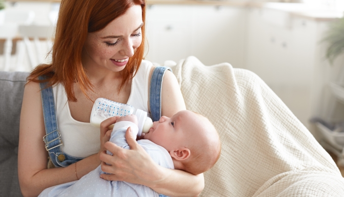Portrait of happy tender young mother with ginger hair feeding her baby boy from milk bottle.