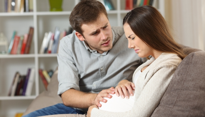 Pregnant woman suffering belly ache and husband comforting her seated on a sofa at home.