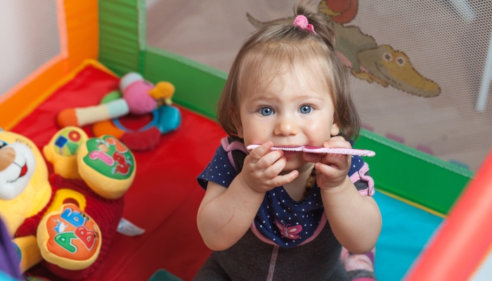 Teething caucasian blonde baby girl playing with a plastic comb in a playard playpen.