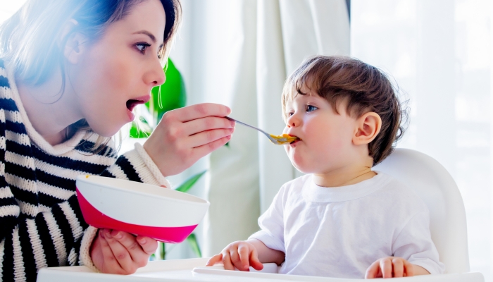 Young mother try to feeding a toddler boy with a spoon in a chair.