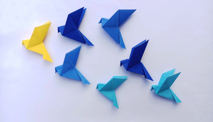Six blue origami birds being led by one yellow origami bird.