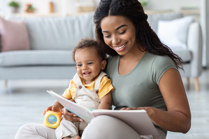 Beautiful Young Black Mommy Reading Book With Her Cute Infant Son At Home.