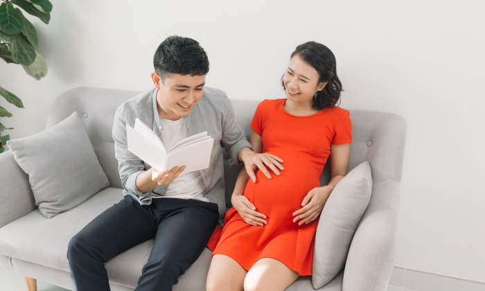 Close-up of a pregnant woman and her husband reading a book on the sofa at home.