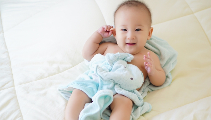 Happy baby wrapped in a towel lying on bed smiling and holding his ear.