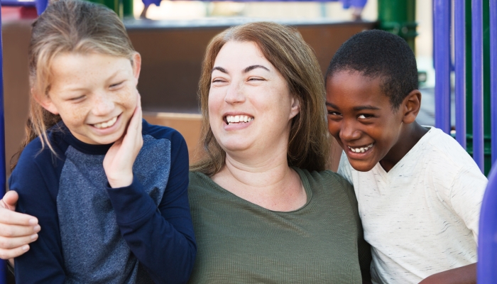 Laughing mother embracing two giggling young sons at park outside.