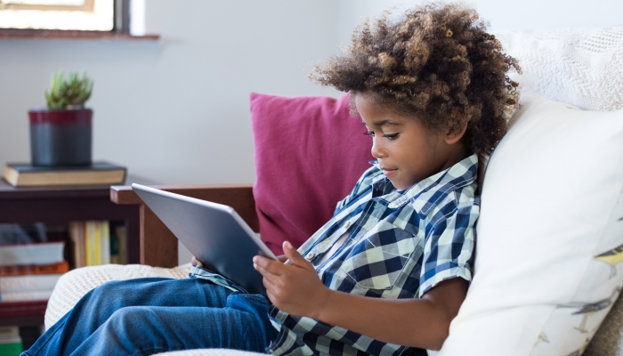Little african boy sitting on sofa and playing game on digital tablet.