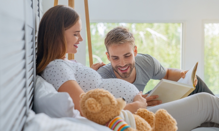 Parents lie on the couch and father read a book to child.