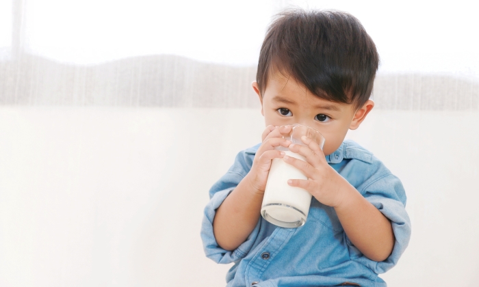 A 1 year and 5 month old boy is sitting drinking milk from a glass.