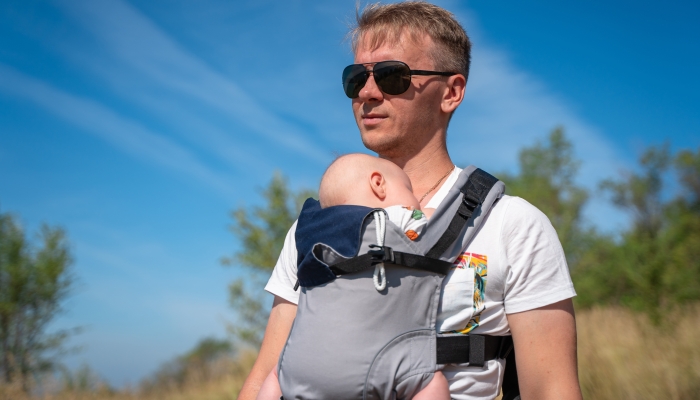 A young father walks with a baby in a sling backpack outside.