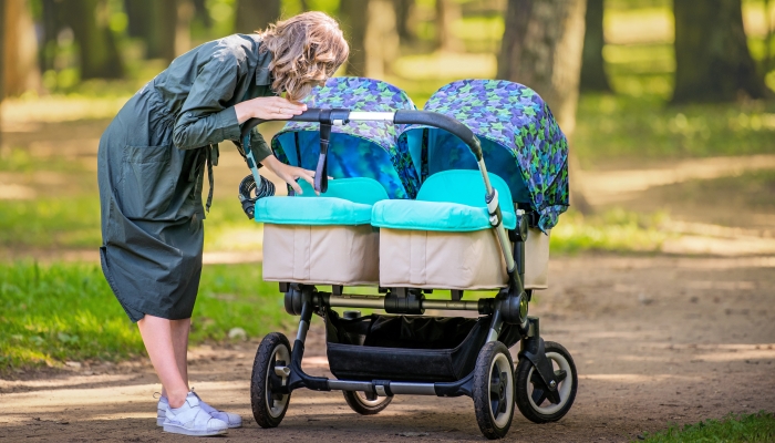 Happy young mom walks in the park in the summer with a stroller for twins, smiling.