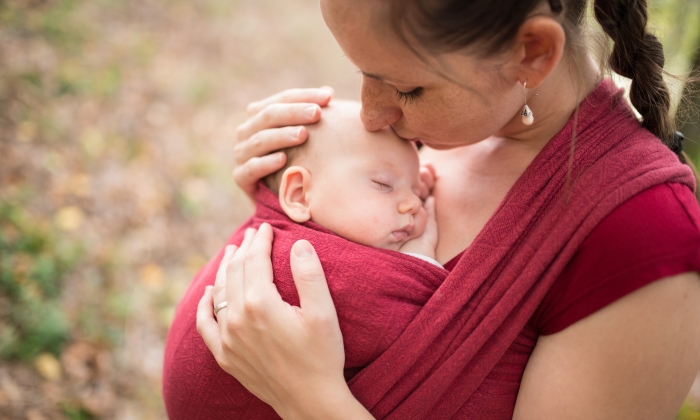 Mother carrying her cute baby daughter in sling, kissing her, outside in autumn nature.