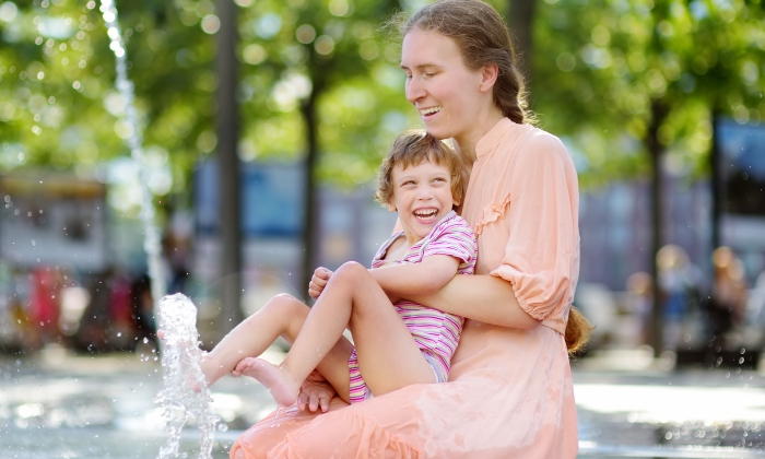 Portrait of beautiful disabled girl in the arms of his mother having fun in fountain of public park at sunny summer day.