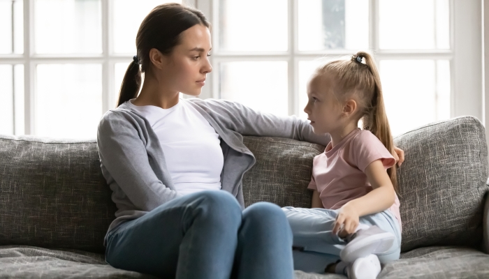 Serious young mom or nanny sit on couch with little preschooler girl talk sharing secrets.