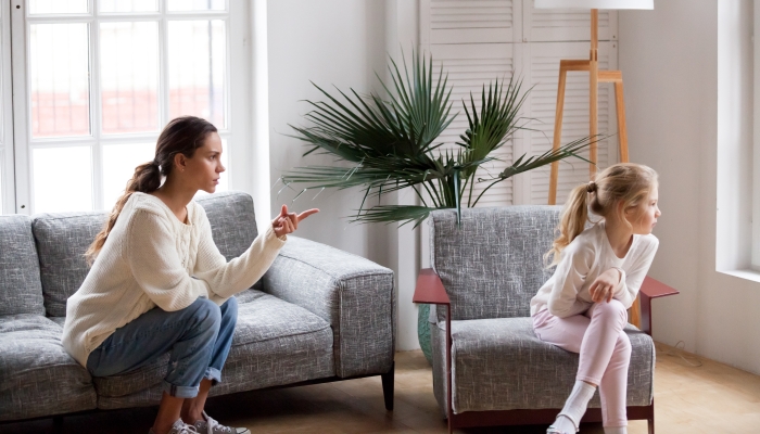 Young strict mother or sister scolding stubborn sulky kid girl in living room at home.