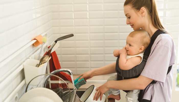 A young mother is washing dishes in the kitchen, the child is fastened to her chest with a baby carrier or babywearing system.