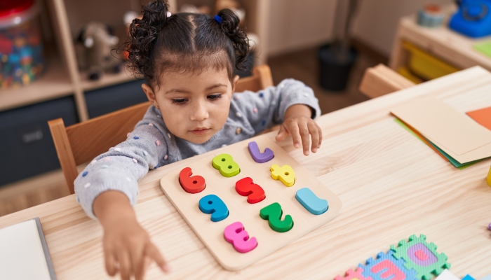 Adorable hispanic girl playing with maths puzzle game sitting on table at kindergarten.