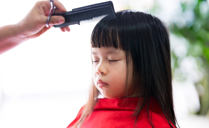Asian kid girl sits still with her eyes closed while hairdresser is styling her hair in order to beautify it.