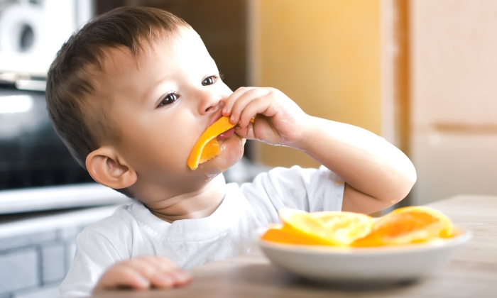 Baby boy in the kitchen eagerly eating an orange.