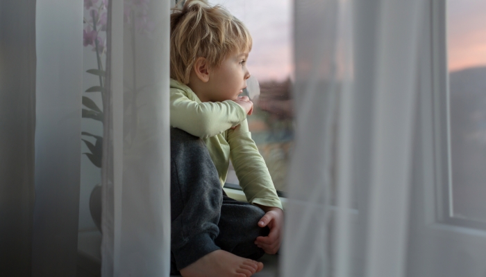 Blond toddler child, sitting on the window on sunset, looking out.