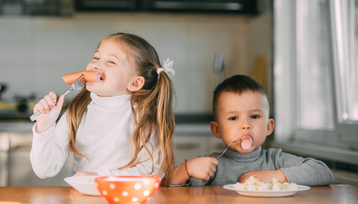 Boy and girl children in the kitchen eating sausages.