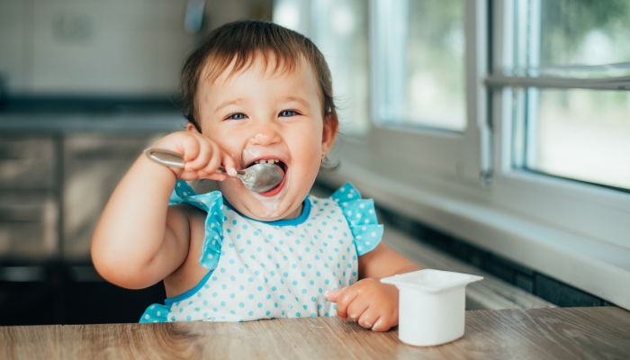 Cute, funny girl eating yogurt during the day in the kitchen in a blue dress.