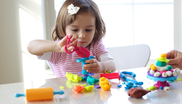 Little girl is learning to use colorful play dough in a well lit room near window.