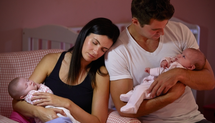 Tired Parents Cuddling Twin Baby Daughters In Nursery.