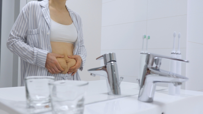 Woman With Postpartum Belly Looking At The Mirror In Bathroom.