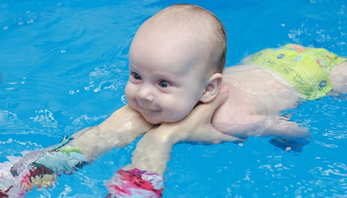 Baby infant learns to swim in the pool, instructor's hands support.