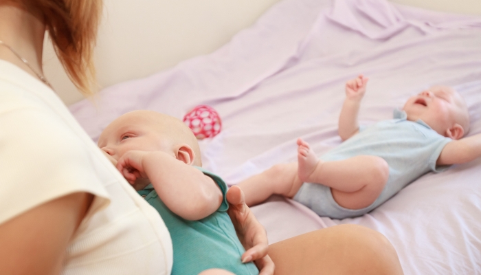 indoor portrait of mother breast feeding and hugging her baby, the other baby is laying next ot her, mother of twin babies at home.