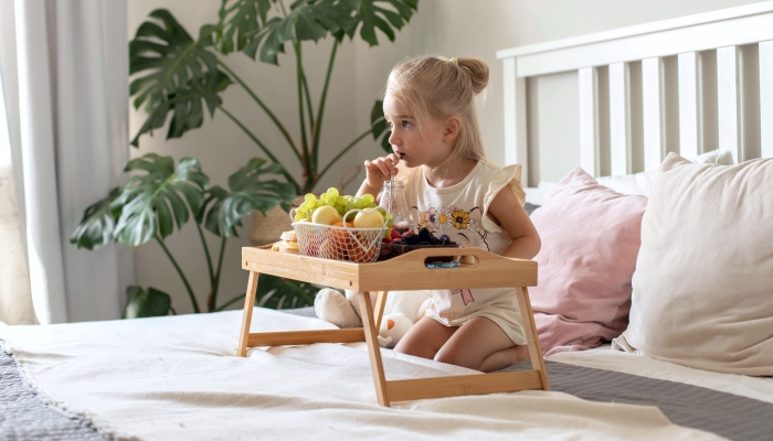 A little girl has breakfast in the bedroom on the bed, drinks smoothies, eats fruits and berries, watches cartoons.