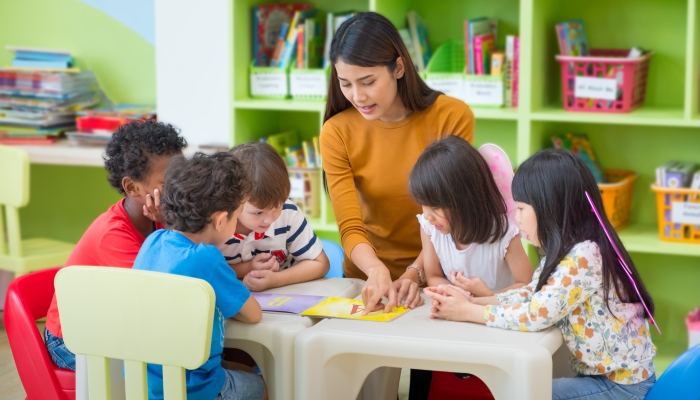 Asian female teacher teaching mixed race kids reading book in classroom.