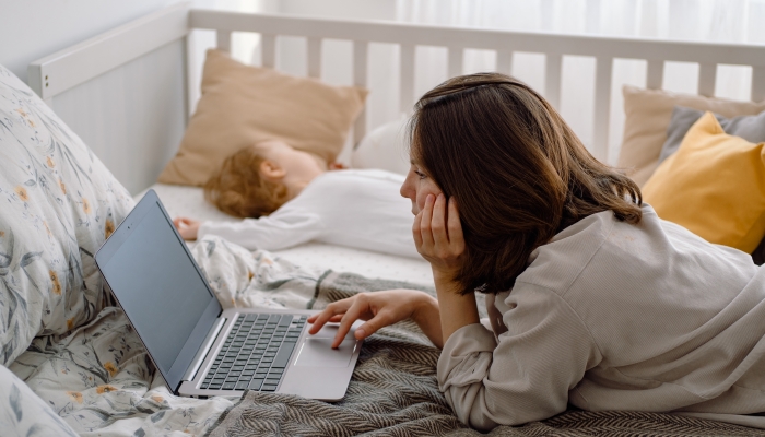 Close up of a young woman mother using laptop lying on the bed, while child sleeps in the crib next to her.