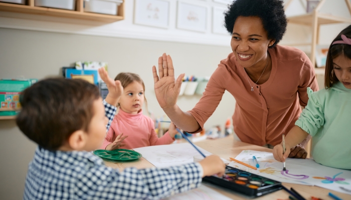 Happy African American teacher and small boy giving high-five during art class at kindergarten.