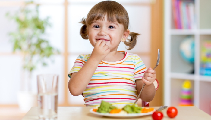 Happy child eats vegetables sitting at table in nursery.