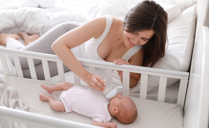 Happy young mother feeding her newborn baby from bottle in bedroom at home.