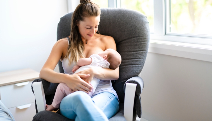 Mother is breastfeeding a baby sitting on a chair close to the window.