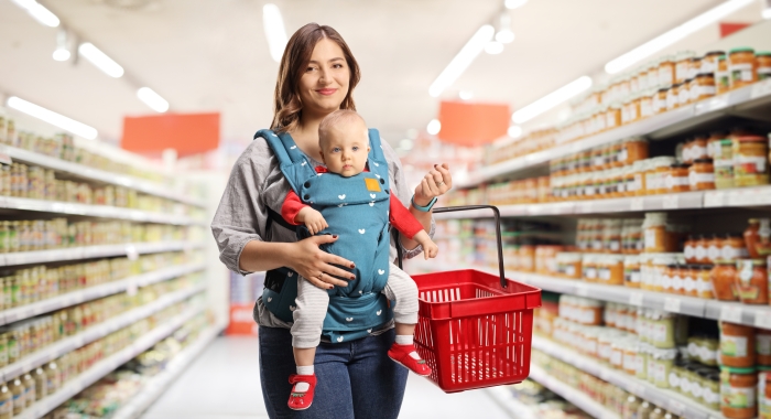 Mother with a baby in a carrier and a shopping basket inside a supermarket.