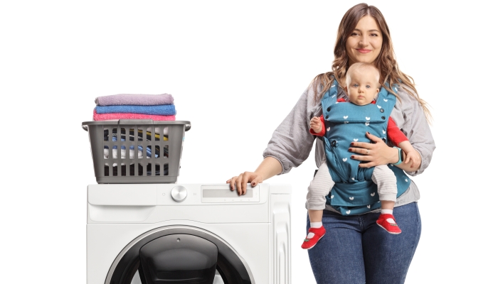 Mother with a baby leaning on a washing machine with a laundry basket.