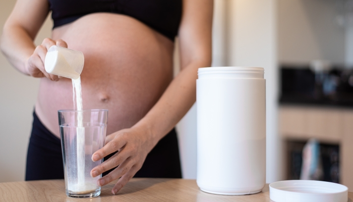 Pregnant woman puring measuring spoon with portion of protein powder into glass.