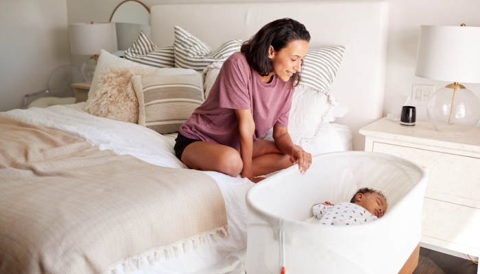 Mother sitting on her bed looking down at her baby sleeping in his cot.