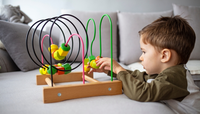 An adorable toddler boy sits at a home in a living room and reaches up cheerfully to play with a toy bead maze.