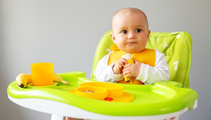 Baby girl eats quinoa porridge and banana while sitting on high chair.