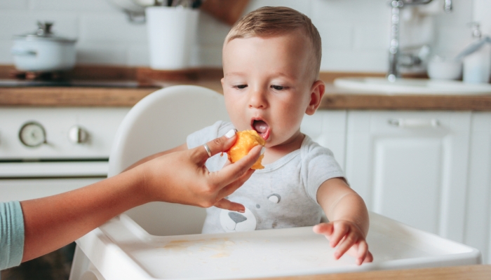 Charming little baby boy eating first food peach at the kitchen.